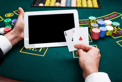 A person holds a pair of playing cards in one hand and a tablet in the other, with poker chips on the table behind it